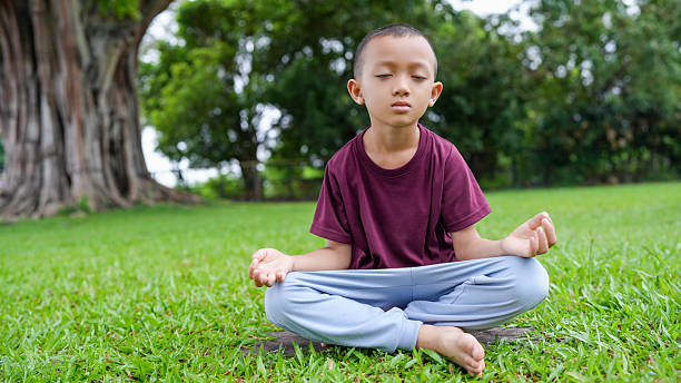 Young boy practices meditation in serene outdoor setting, surrounded by lush greenery and large tree. His calm expression reflects tranquility and focus, embodying peaceful moment of mindfulness