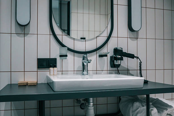 istock Modern hotel bathroom interior featuring oval mirror, white-tiled walls, sleek countertop basin, chrome faucet, wall-mounted hairdryer and neatly rolled towels for a spa-like ambience 2229508612