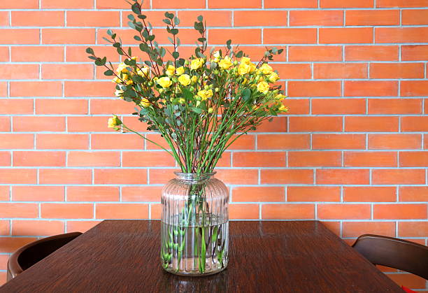 Big glass bottle with bright yellow rose in on dark brown wood table and red brick wall background.