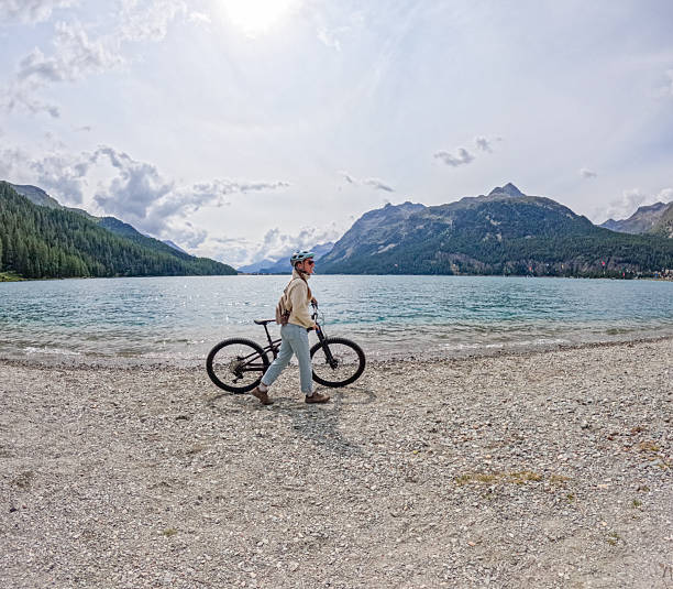 Young woman wearing helmet and backpack stands with bicycle on rocky shore by mountain lake. Outdoor adventure and cycling lifestyle in nature. Scenic landscape with water, forest, and mountains.