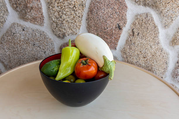 A bowl of vegetables including tomatoes, peppers, and squash. The bowl is black and sits on a wooden table