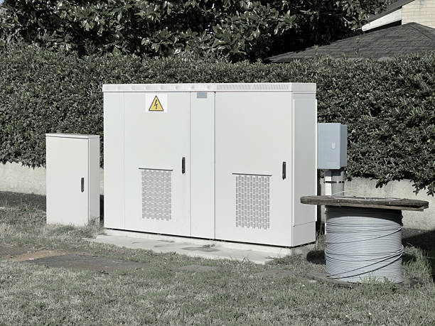 A large white roadside utility cabinet with a yellow high-voltage warning symbol stands on a concrete pad beside a smaller service box and a meter enclosure. A wooden reel holding gray cable sits on the grass verge in front. A hedge and nearby houses form the background. Photographed on a residential street in Italy; equipment appears suited for electrical or telecommunications distribution.