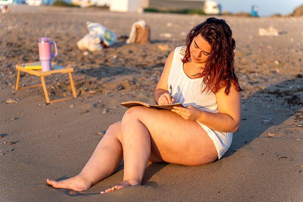 istock Curvy woman writing on notebook at sunset on the beach 2229522484