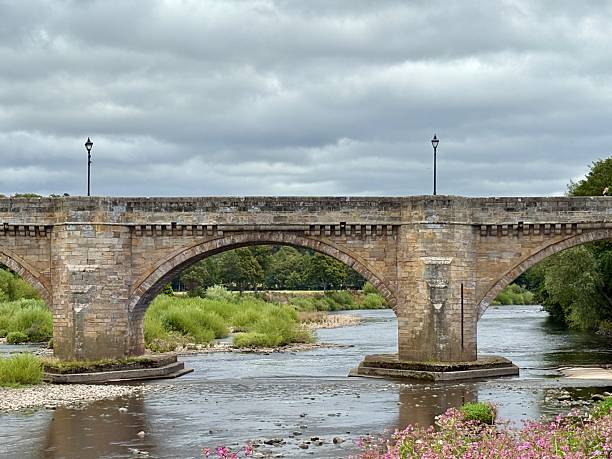 istock A historic stone bridge spans a calm river beneath a cloudy sky, with wildflowers blooming in the foreground. This picturesque scene blends architecture and nature in a peaceful countryside setting. 2229525464
