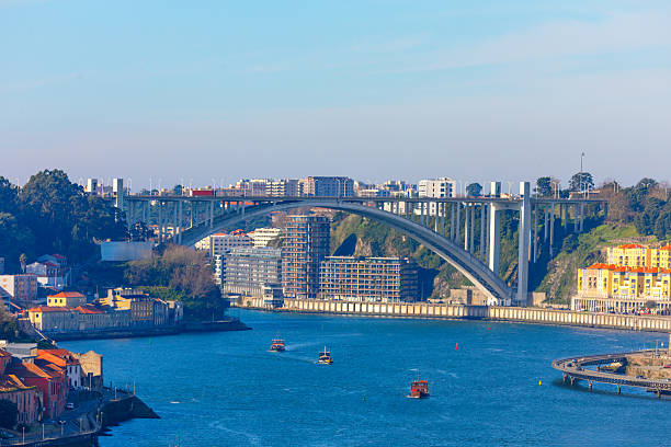 Elegant Ponte da Arrabida spans the Douro River in Porto, framed by colorful riverside buildings and modern apartments. Small boats glide beneath sweeping arch on sunny day
