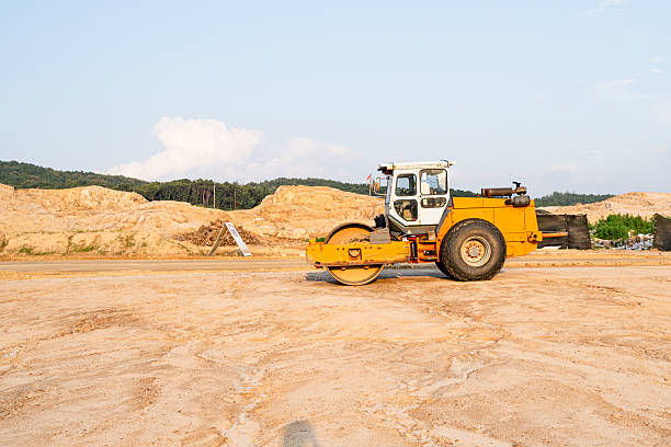 A bright yellow road roller compacts soil at a construction site, surrounded by dirt mounds and distant greenery, preparing the ground for upcoming roadwork and infrastructure development.