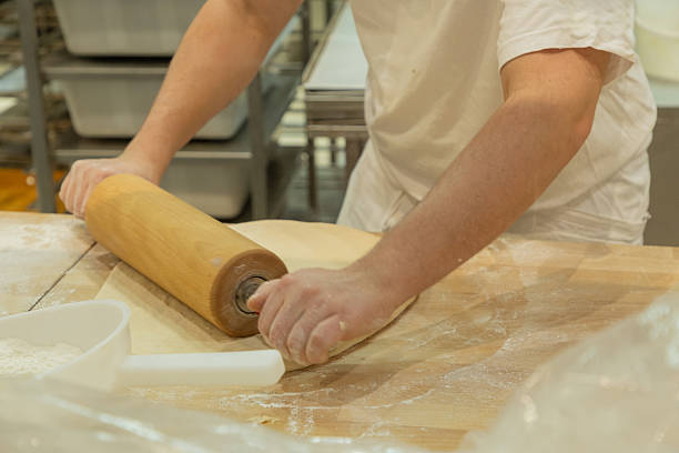 istock Baker rolling dough on wooden surface in a professional kitchen with flour scattered around 2229536048