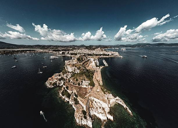 An aerial view of a coastal town with historic fortifications and boats on a sunny day, showcasing vibrant blue waters and dramatic clouds.