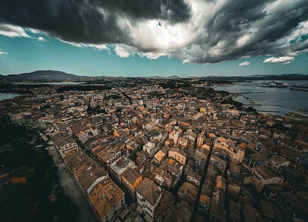 An aerial view of Corfu town under dramatic clouds with a cruise ship in the distance, showcasing the city's dense architecture and coastal beauty.
