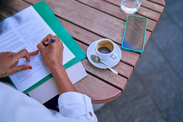 Close-up of an Asian woman seated at a table, focused on her work. She is holding pens and a personal organizer, diligently making notes in documents spread out in front of her. In the midst of her tasks, she enjoys a cup of coffee, creating a balanced and productive workspace