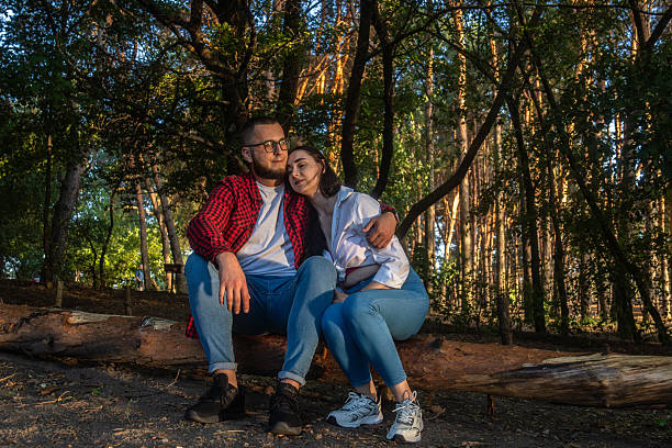 Young couple sitting next to each other on a fallen tree trunk in the forest during golden hour. Romantic man and woman enjoying a peaceful and intimate moment surrounded by nature.