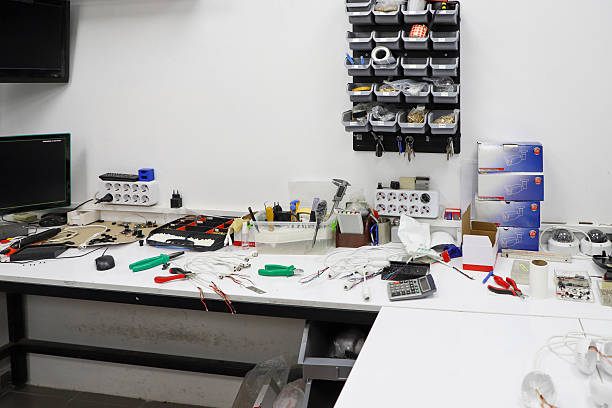 An image of a cluttered workshop or electronics lab. In the foreground is a workbench with various tools, cables and electronic components strewn about.