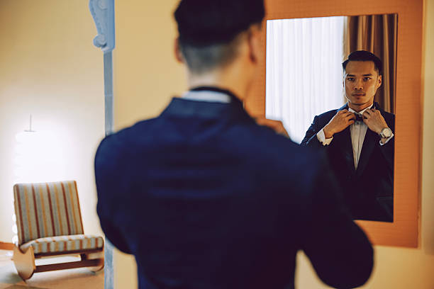 Chinese man in a dark suit and bow tie adjusts his bow tie while looking into a mirror in a warmly lit hotel room, preparing for a formal event, Kuala Lumpur, Malaysia.