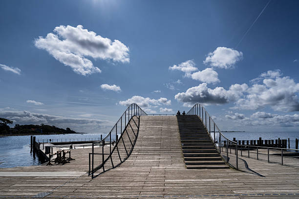 A scenic view featuring a distinctive wooden pier with stairs designed in a wave-like shape, surrounded by calm water and a clear blue sky, evoking feelings of tranquility and innovation.