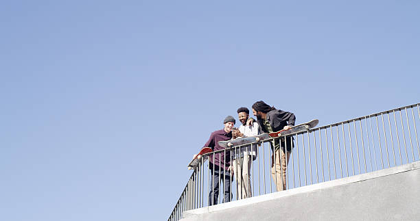 Below, people and laughing with skateboard, phone and meme together on blue sky space. Bonding, friends and gen z skater group with tech, smile and social media for fitness hobby and break for sport