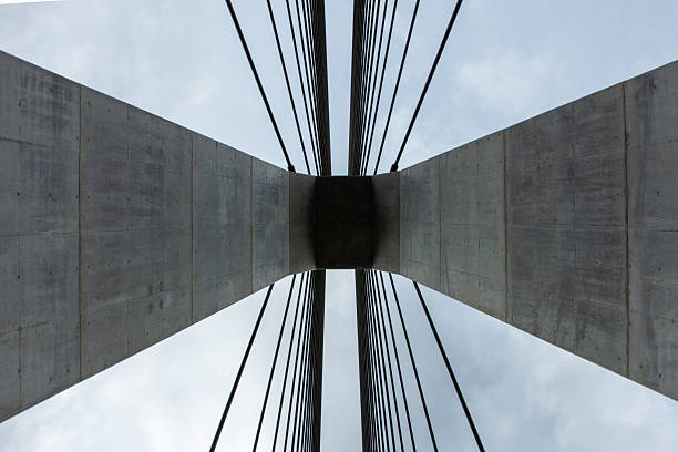 An abstract, symmetrical, low-angle photograph looking up at the main pylon of the Gakkaku Sazanami Ohashi, a modern cable-stayed bridge in Japan. The powerful concrete structure and the fanned-out steel cables create a striking geometric pattern against the overcast sky. This image is perfect for themes related to modern architecture, civil engineering, construction, technology, and infrastructure.