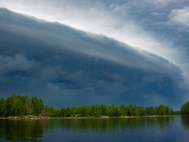 This breathtaking and ominous image captures the exact moment a powerful shelf cloud dominates the sky, signaling the arrival of a severe storm. The cloud formation stretches from left to right in dramatic bands, rolling low and heavy over a peaceful forest-lined lake. The contrast between the dark, brooding clouds and the calm, reflective water below creates a visually stunning tension. The scene is remote, wild, and untouched—an intense display of nature’s duality between calm and chaos. The pine trees along the shore are sharply defined, anchoring the frame and enhancing the vastness of the cloud system looming above.