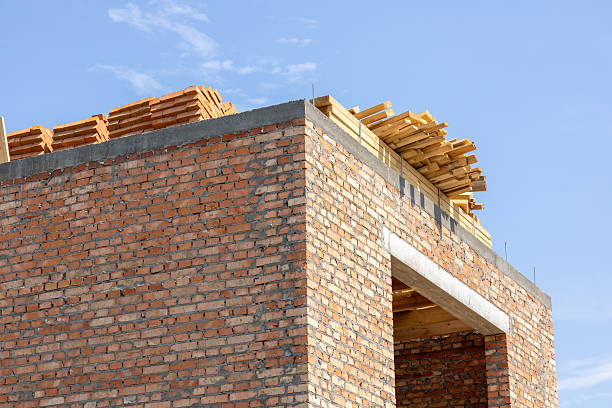 Workers are busy on a construction site where a brick building is being erected, showcasing wooden planks and bricks against a clear sky.
