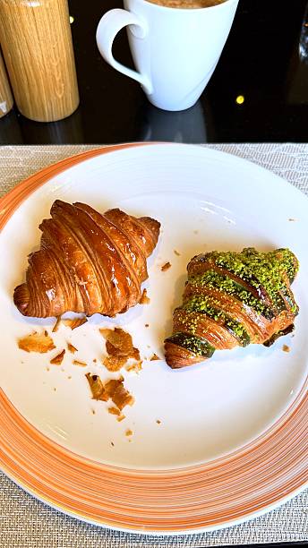 Plain croissant and pistachio croissant served on plate for breakfast, accompanied by cappuccino, breakfast offering delightful flavors and inviting aroma for perfect start to day.