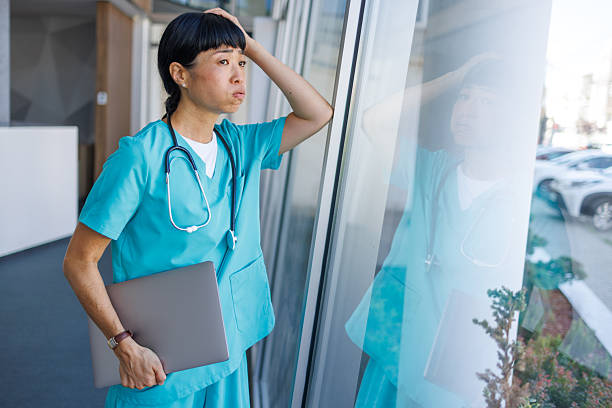 istock Overworked nurse leaning against window holding laptop 2229583607