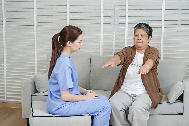 istock Senior asian woman lifting both arms slowly forward while sitting on sofa in rehab session guided by young female therapist wearing blue uniform observing carefully in clinic 2229583902