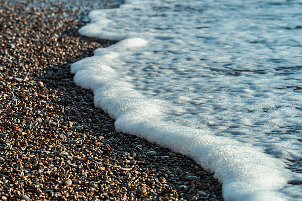 White sea wave foam washing over colorful pebble beach. Natural seascape showing the meeting point of sea water and rocky coastline with smooth rounded stones.