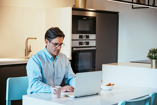 istock Motivated businessman sitting at a kitchen table working on a laptop, illuminated by warm light, with a smartphone and plant nearby 2229585429