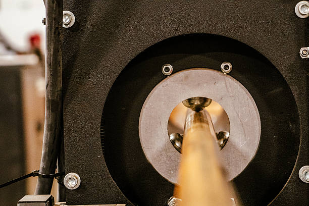 A close-up image of a cylindrical metal rod or tube being fed through a circular opening in a black industrial forming or testing machine. The focus is on the mouth of the machine, where polished metallic guides or rollers stabilize the material. The environment appears to be an indoor manufacturing or mechanical engineering facility, with visible bolts and wiring on the frame of the equipment. The blurred foreground suggests a fast-moving or continuous process, emphasizing industrial precision and mechanical function.