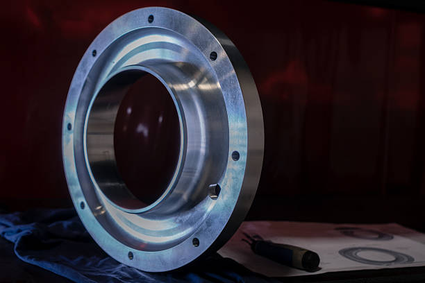 A precisely CNC-machined flanged metal ring sits upright on a blue inspection cloth next to technical engineering drawings and a handled deburring tool. The part features multiple bolt holes around the flange and a clean, smooth internal surface, indicating precision tolerances. The setup rests on a dark workbench labeled \
