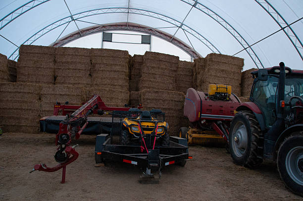 Old red farm tractor and cultivator outside of a barn and tall grain silos on a farm