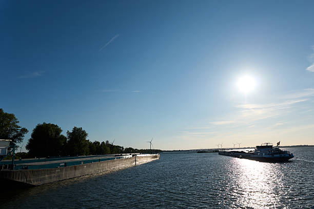 Barge sailing on a river on a sunny day, with wind turbines and trees on the shore