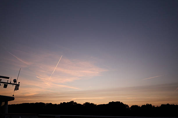 Airplane is flying over the river at sunset, leaving a white contrail in the colorful sky