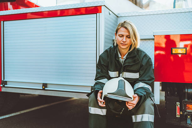 A mature Caucasian male airport firefighter in black and yellow protective gear climbs into a bright red fire truck. The well-lit scene highlights the vehicle's bold colors and functional design during a professional emergency response setting.