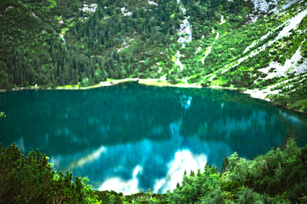Mountain lake with a deep blue water reflecting clouds, surrounded by green forested slopes .