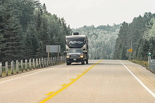 Algonquin Park, Ontario, Canada, July 30, 2025. Vehicles driving along a winding, hilly stretch of Highway 60 as it passes through dense forest in Algonquin Provincial Park, Ontario, Canada. The road climbs gently toward the horizon, bordered by tall trees on both sides, creating a strong sense of depth and movement. Soft, natural lighting from early morning or late afternoon adds a peaceful ambiance to the scene. Clear road markings include solid yellow center lines and dashed white edge lines. Roadside signs are visible, providing important information to travelers navigating this scenic route.