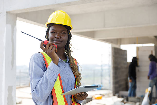 istock Female african american construction engineer using walkie-talkie and holding tablet while supervising site progress. Wearing safety vest and yellow hardhat, she represents communication. 2229652061