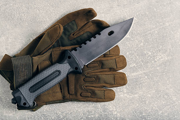Military tactical knife lying on gloves, gray concrete background.