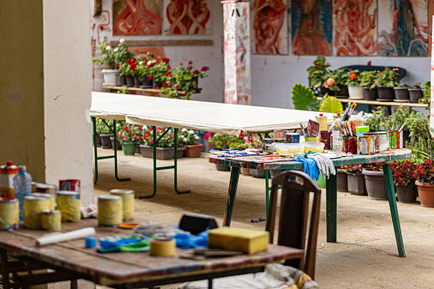 An outdoor art studio scene shows a table with paints, brushes, and tools in the foreground, with a prepared canvas on another table in the background, ready for artistic creation.
