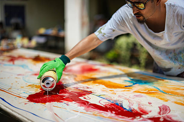A sunlit shot captures a male artist wearing sunglasses and protective gloves, carefully applying solvent to his vibrant abstract painting on a large canvas outdoors.