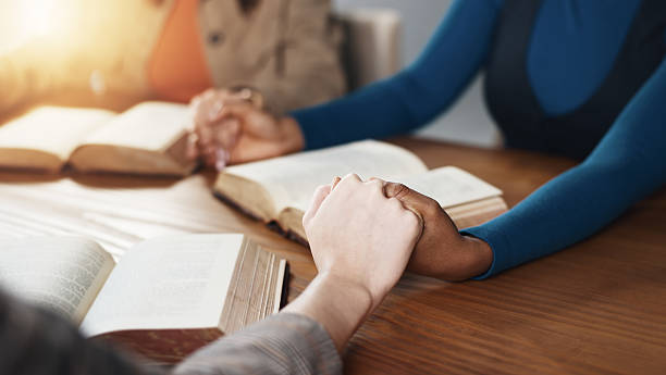 istock Hand holding, Bible and people praying with religion, guidance or support with scripture for worship. Christianity, faith and group together by table, book of God and praise with spiritual education 2229683478