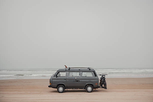 a classic dark gray camper van parked on a sandy beach, facing the ocean under an overcast sky. A surfboard fin sticks out from the roof rack, and a motorcycle is mounted on a rear carrier, suggesting a spirit of adventure and multi-sport travel. The waves roll gently in the background, and the muted tones of the scene give it a calm, minimalist vibe that blends surf culture with van life freedom.