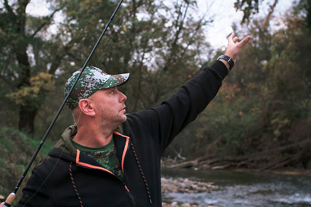 A fisherman in camouflage attire fishing by a serene stream surrounded by nature. Man tries to replace the bait on the hook on the fishing rod. The tranquil outdoor scene showcases the hobby of fishing and the beauty of wilderness environments