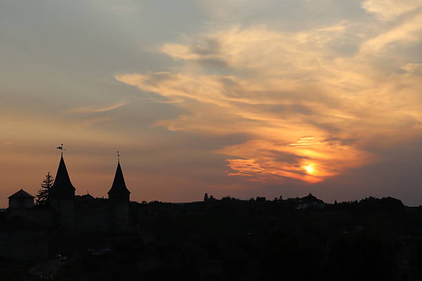 The sun sets behind a historic castle, casting a warm glow across the sky. The silhouettes of towers stand prominently against the vibrant colors, creating a serene atmosphere.