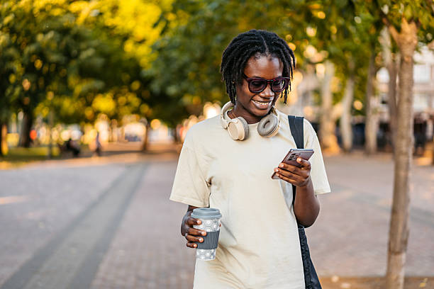istock Young Black Woman Using Phone And Drinking Coffee While Walking In The Public Park 2229721642