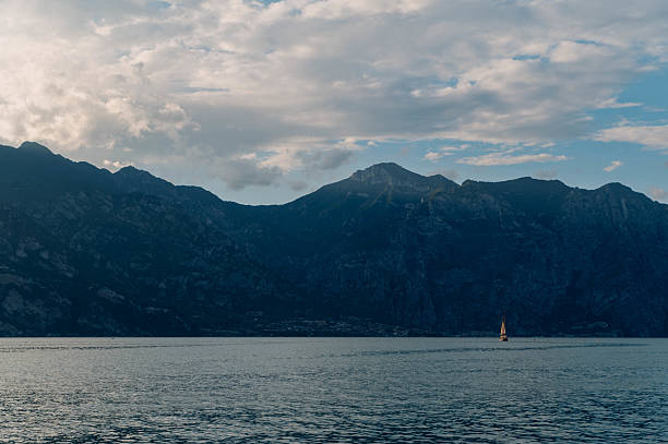 A horizontal view of Lake Garda featuring mountains in the background and the silhouette of a sailboat far across the water.