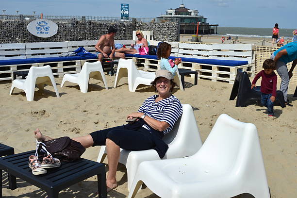 Blankenberge, West-Flanders, Belgium- August 3, 2025: happy woman in her early sixties, wearing a cap, sits on a plastic beach chair at a surf beach club, smiling. A curious attentive blonde woman in background