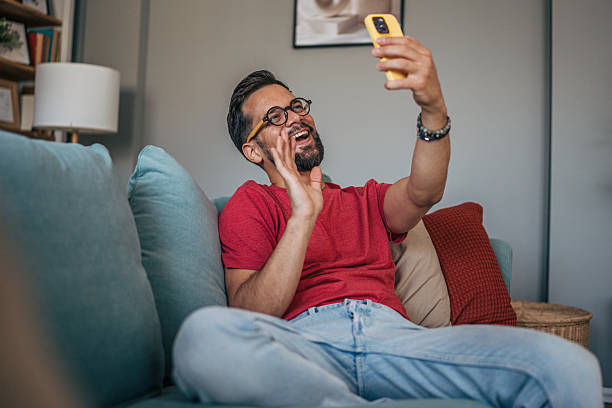 istock Happy man video calling with friends while relaxing on sofa 2229750955