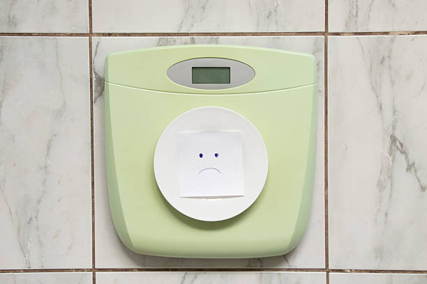 Green electronic floor scales on white tile with empty white plate and paper with sad face, diet and food restriction concept