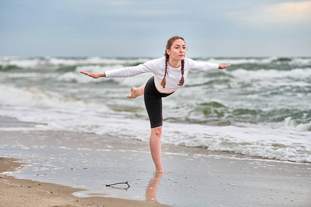 Young woman with braided hair practices yoga on baltic sea beach. Her focused pose contrasts with serene waves and cloudy sky. Evening light casts soft shadows on sand