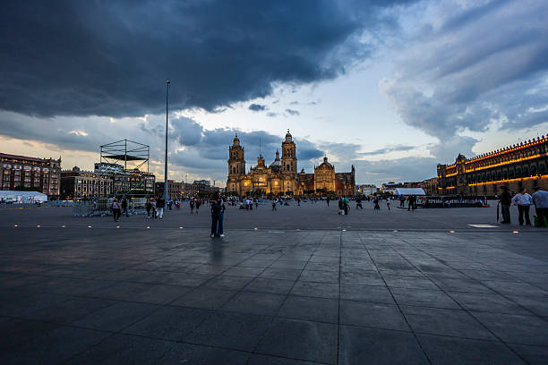 Mexico City, Mexico, July 3rd, 2025. Zocalo Square in Mexico City with the Metropolitan Cathedral and National Palace illuminated at twilight with storm clouds and visitors walking around it.